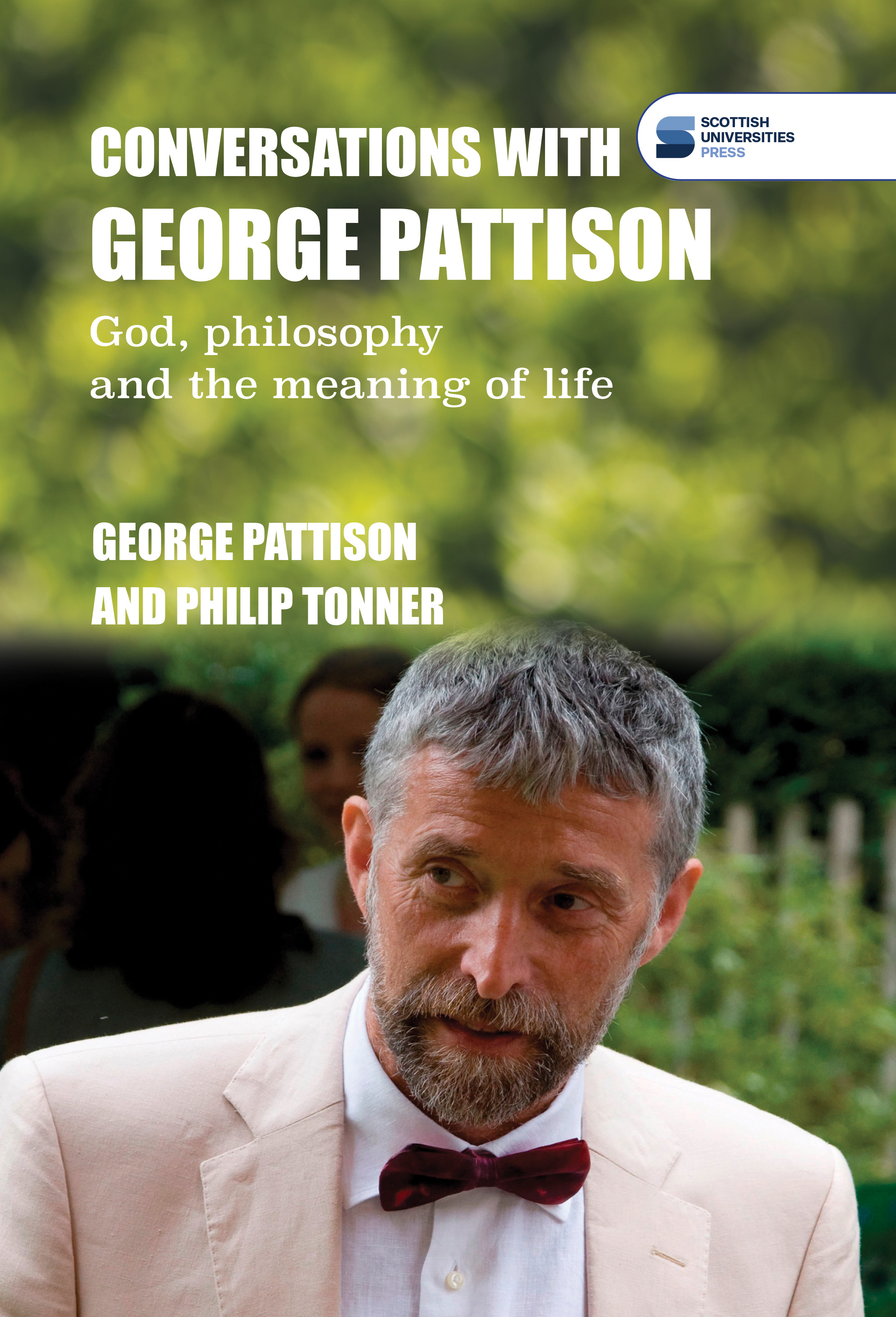 Photo of George Pattison wearing a light suit and red bowtie, with a blurred background of trees and other greenery.